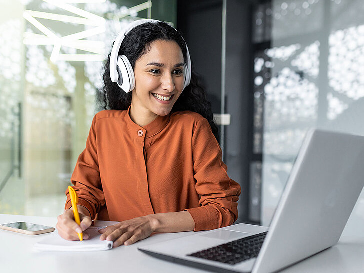 Latin american business woman with curly hair and headphones watching online training course at workplace, woman writing information happy and satisfied with the results of professional development